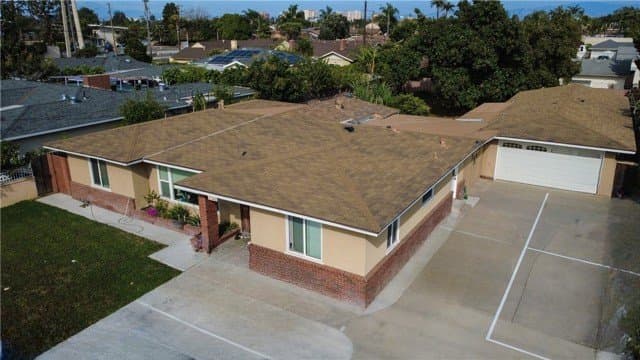 Aerial view of a single-story house with a brown shingle roof, tan stucco walls, and a red brick lower facade, a large concrete driveway, and a front lawn.