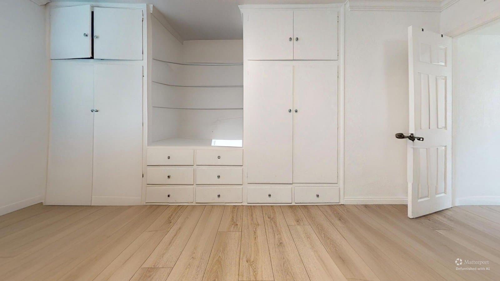 Virtual rendering: An empty room with light wood floors, white built-in cabinets and shelves along the walls, and a partially open white door on the right.